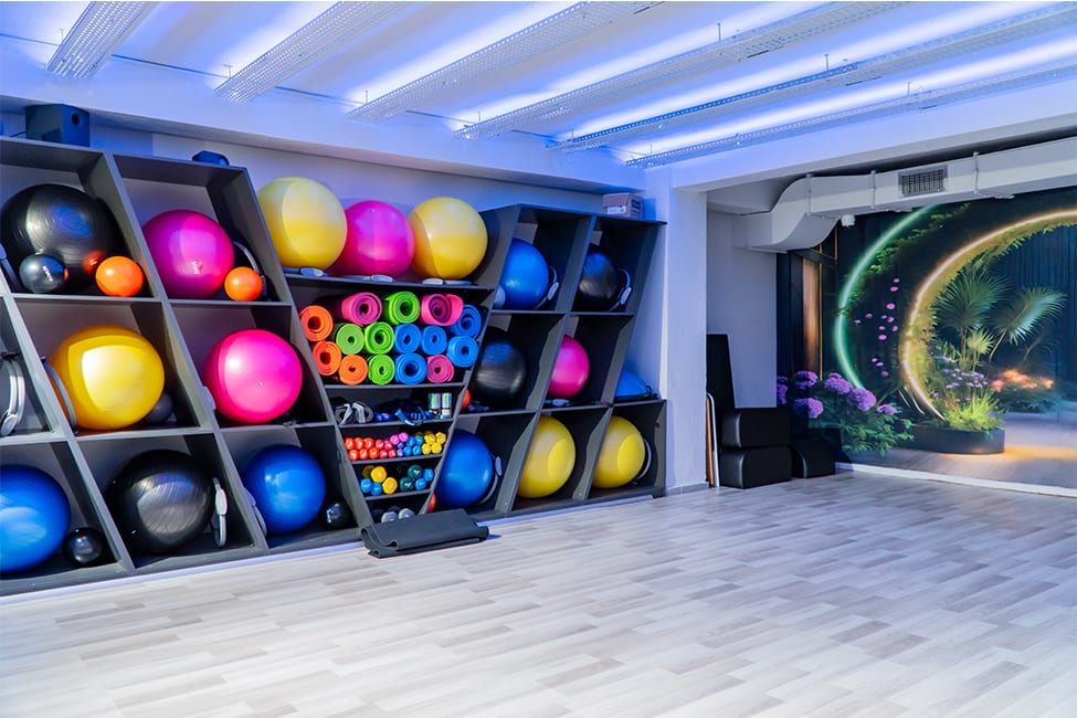 Today Living Club grup dersi stüdyosu: raflarda renkli pilates topları, yoga matları ve dambıllar, duvarda neon kemerli bahçe görseli